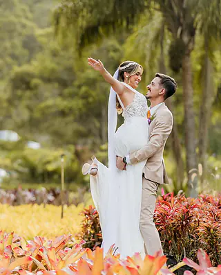 Noivo levanta a noiva no colo num jardim tropical, ela sorri com o braço aberto