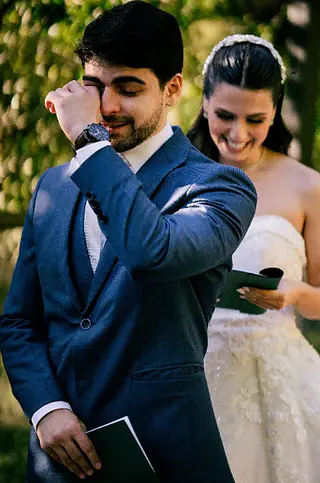 Noivo enxuga as lágrimas segurando uma carta, com a noiva sorrindo logo atrás