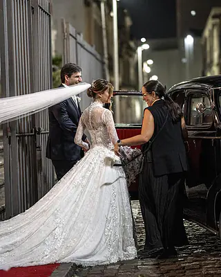 À noite, a cerimonialista ajuda a noiva a chegar ao carro antigo enquanto o noivo sorri